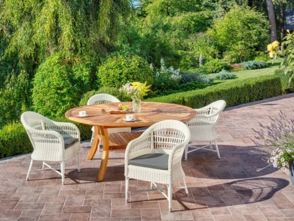 Round teak garden table with white wicker armchairs among greenery