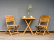 Două scaune pliante Ambra și o măsuță pătrată din tec cu căni de cafea în fața unui perete gri